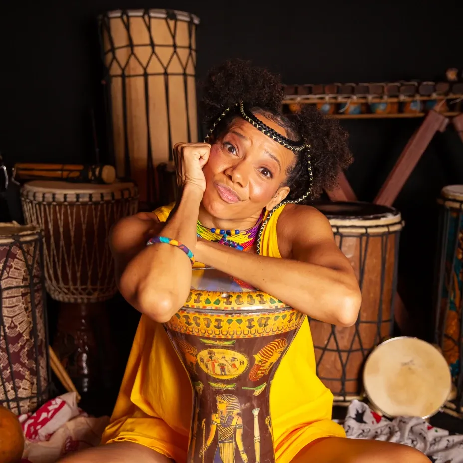Akili sentada apoiada em tambor decorado, vestindo roupa amarela e acessórios étnicos coloridos, rodeada por djembês e instrumentos de percussão africanos em fundo escuro.