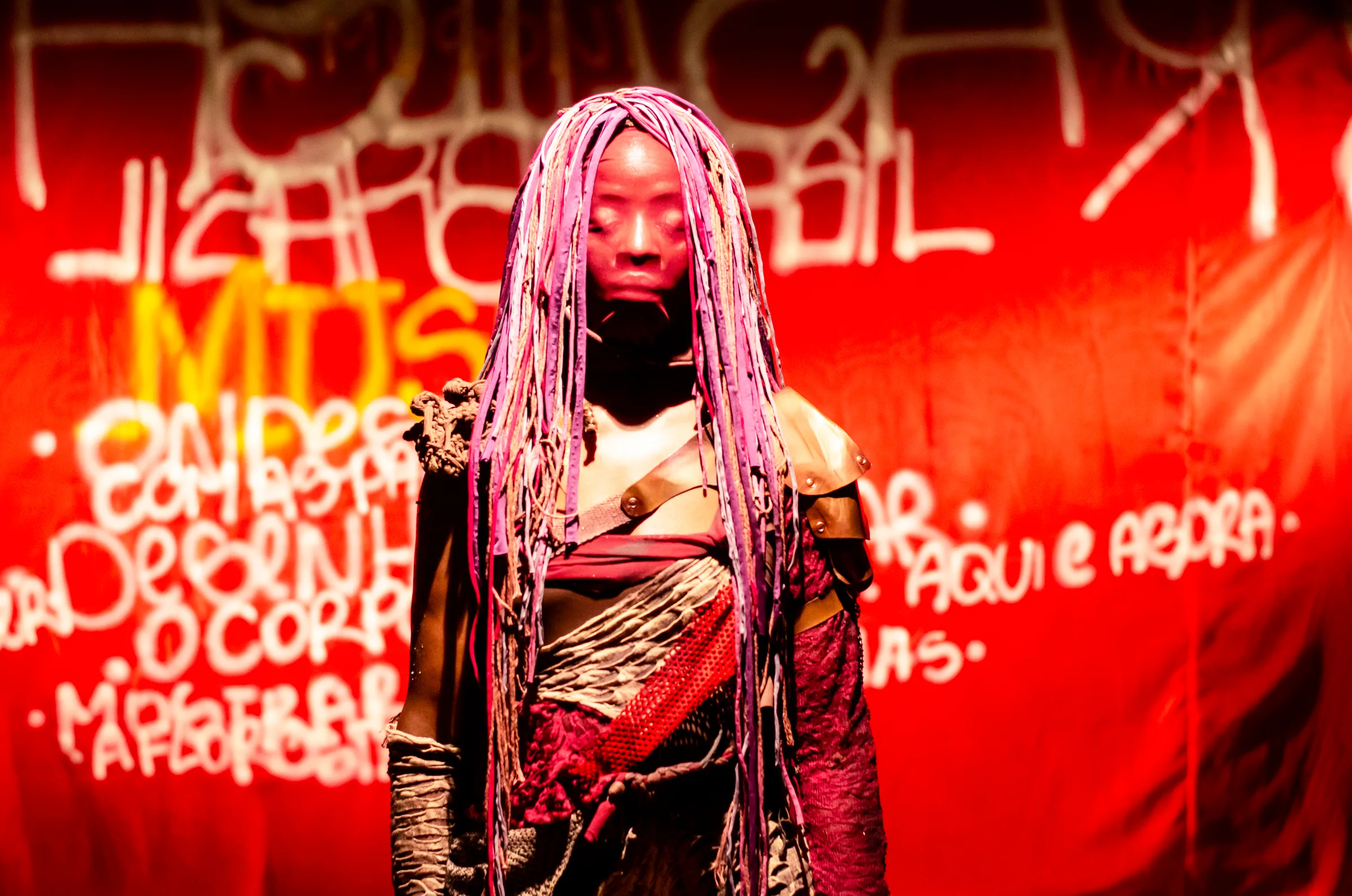 Atriz em cena teatral com figurino experimental em tons de vermelho e roxo, incluindo longas tranças de fios, diante de fundo vermelho com inscrições pintadas, sob iluminação intensa.